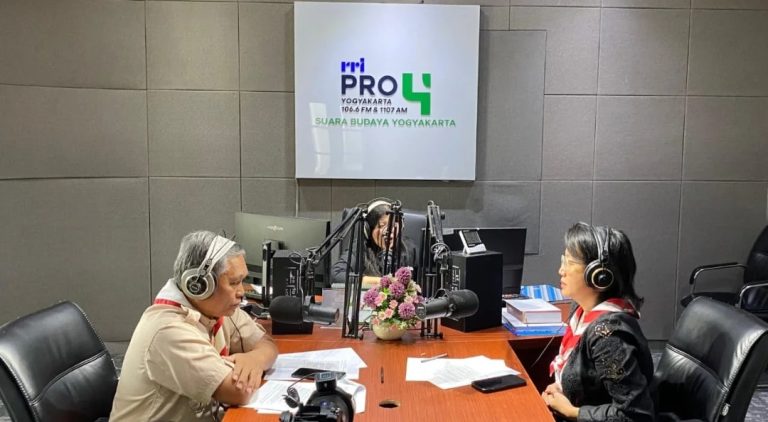 Three radio hosts wearing headphones sit around a wooden table with multiple microphones in a studio, with a logo wall behind them.
